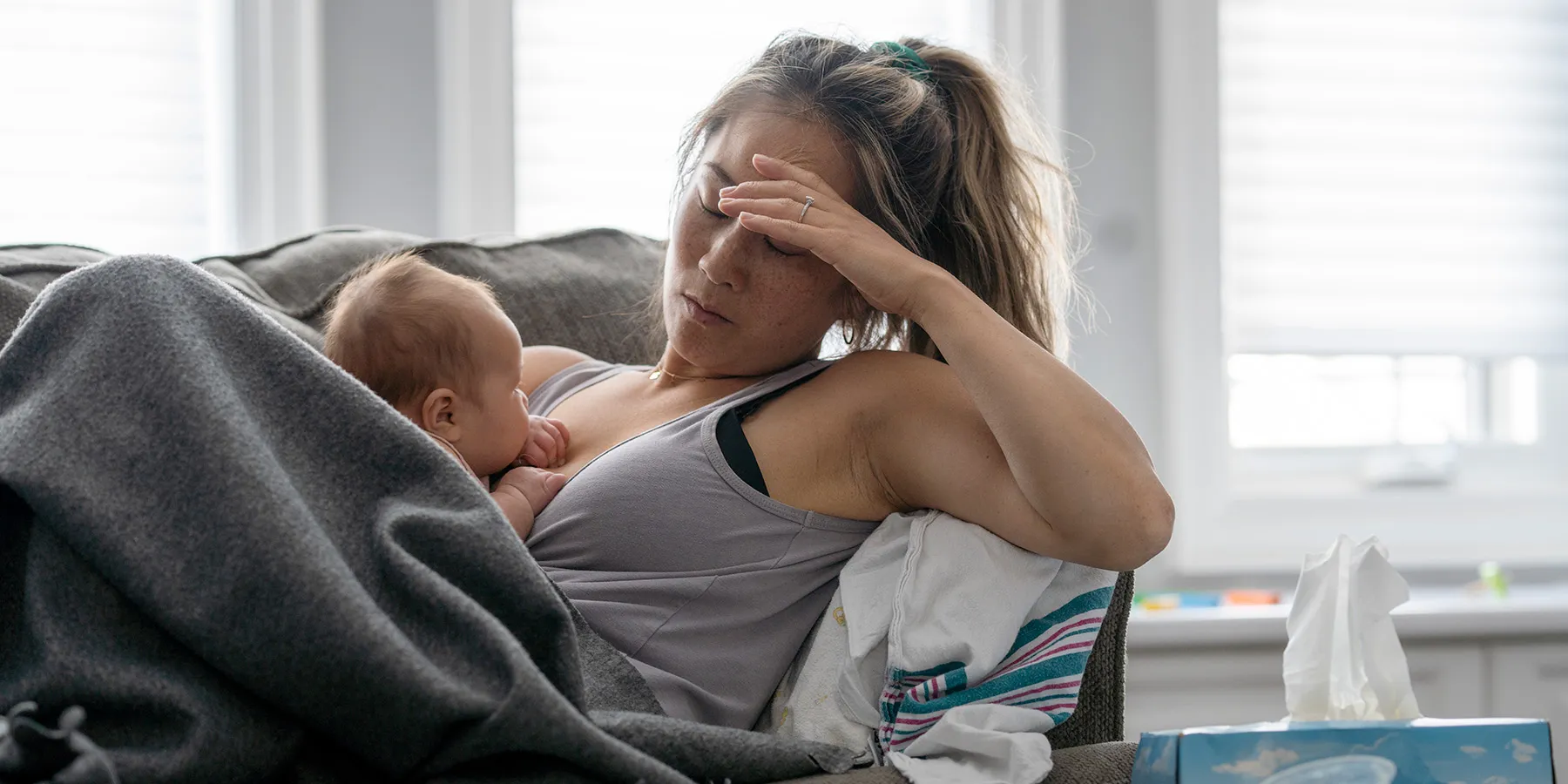 A middle-aged mother resting on her couch holds her baby in her arm and holds her other hand over her head with a tired and stressed expression.
