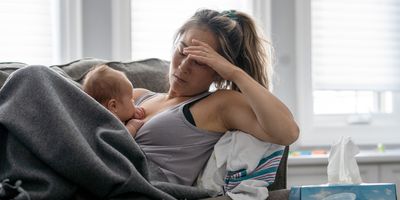 A middle-aged mother resting on her couch holds her baby in her arm and holds her other hand over her head with a tired and stressed expression.