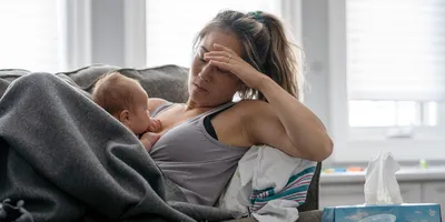 A middle-aged mother resting on her couch holds her baby in her arm and holds her other hand over her head with a tired and stressed expression.