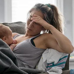 A middle-aged mother resting on her couch holds her baby in her arm and holds her other hand over her head with a tired and stressed expression.