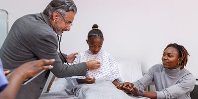 A sick young girl sits in a hospital bed as her doctor uses a stethoscope to check her heart and lungs. The child's mother is sitting next to the bed and is holding her daughter's hand, providing support and comfort.