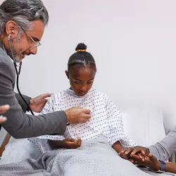 A sick young girl sits in a hospital bed as her doctor uses a stethoscope to check her heart and lungs. The child's mother is sitting next to the bed and is holding her daughter's hand, providing support and comfort.