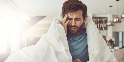 A man having a headache touches his right temple as he sits wrapped in a duvet on a bed.