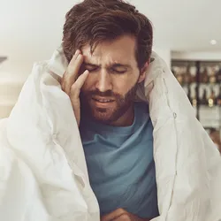 A man having a headache touches his right temple as he sits wrapped in a duvet on a bed.