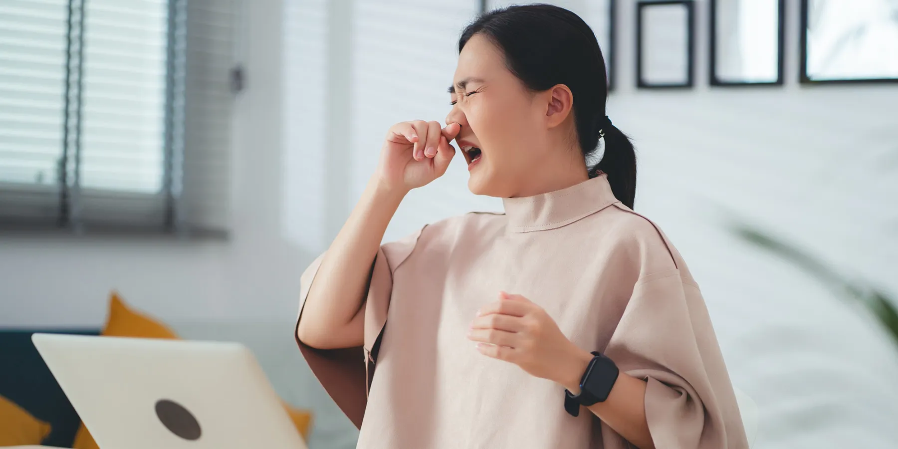 A woman sitting at her desk with laptop, phone, etc., rubs her nose with her right and touches her throat with her left hand to indicate infection.