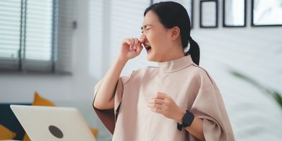 A woman sitting at her desk with laptop, phone, etc., rubs her nose with her right and touches her throat with her left hand to indicate infection.