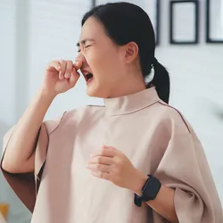 A woman sitting at her desk with laptop, phone, etc., rubs her nose with her right and touches her throat with her left hand to indicate infection.