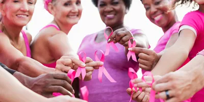 A multiethnic group of five women of mixed ages stand together outdoors, each holding a breast cancer awareness ribbon near the camera.