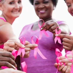 A multiethnic group of five women of mixed ages stand together outdoors, each holding a breast cancer awareness ribbon near the camera.