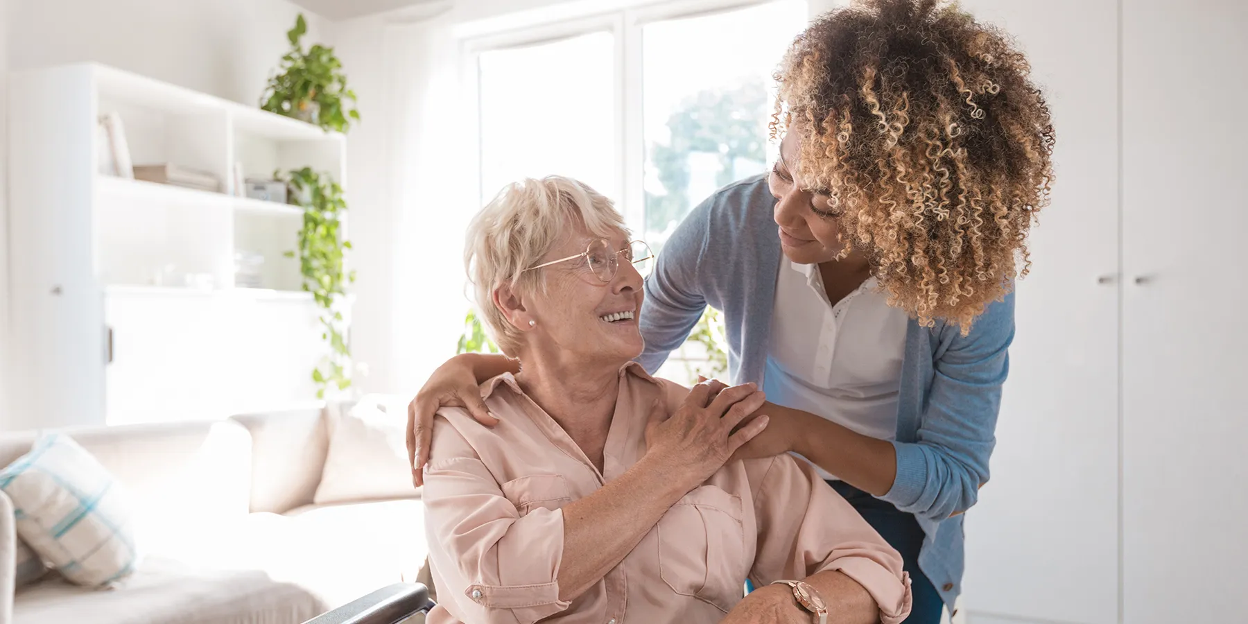 An elder woman in a wheelchair with Parkinson's smiles at a cheerful personal care worker who's crouching over the wheelchair, resting her hands on the elder woman.