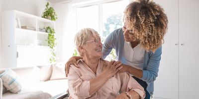 An elder woman in a wheelchair with Parkinson's smiles at a cheerful personal care worker who's crouching over the wheelchair, resting her hands on the elder woman.