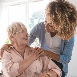 An elder woman in a wheelchair with Parkinson's smiles at a cheerful personal care worker who's crouching over the wheelchair, resting her hands on the elder woman.
