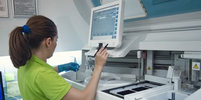 A female researcher works on a large instrument as she holds a sample tube with a gloved left hand.