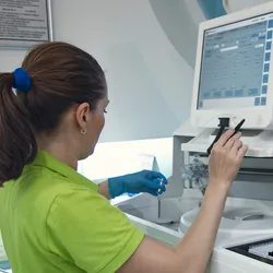 A female researcher works on a large instrument as she holds a sample tube with a gloved left hand.