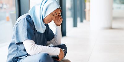A burnt-out female healthcare worker dressed in scrubs and a headscarf sits cross-legged on the floor with her left hand holding her head in stress.