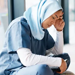A burnt-out female healthcare worker dressed in scrubs and a headscarf sits cross-legged on the floor with her left hand holding her head in stress.