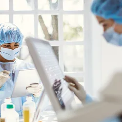 Two female researchers in PPE work on digital tablet and touch-screen computer interface in a  lab setting.