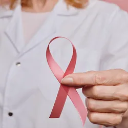 A female clinician holds a pink ribbon in front of the lens symbolizing breast cancer awareness.