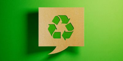 Photo of a chat bubble made from cardboard with a cut-out in the shape of a recycling symbols with three arrows on a green background.