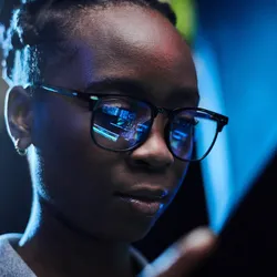 Photo of a young Black female artificial intelligence engineer using a digital tablet while working in a dark computer server room.