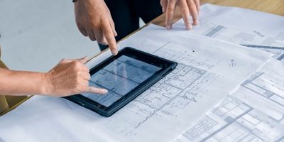 Photo of two project managers standing over a table with lab blueprints on it and pointing at a table with designs on it.