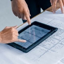 Photo of two project managers standing over a table with lab blueprints on it and pointing at a table with designs on it.