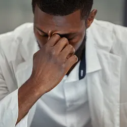 Photo of a Black medical laboratory scientist wearing a white lab coat resting his head on his hand and looking down at the laboratory bench.