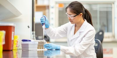 Photo of a female medical laboratory scientist wearing PPE and sitting at the lab bench pipetting.