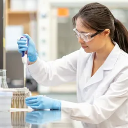 Photo of a female medical laboratory scientist wearing PPE and sitting at the lab bench pipetting.