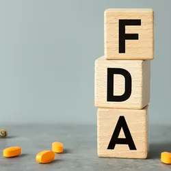 Photo of the letters F, D, and A, representing the US FDA, on wooden blocks with pills in the background.