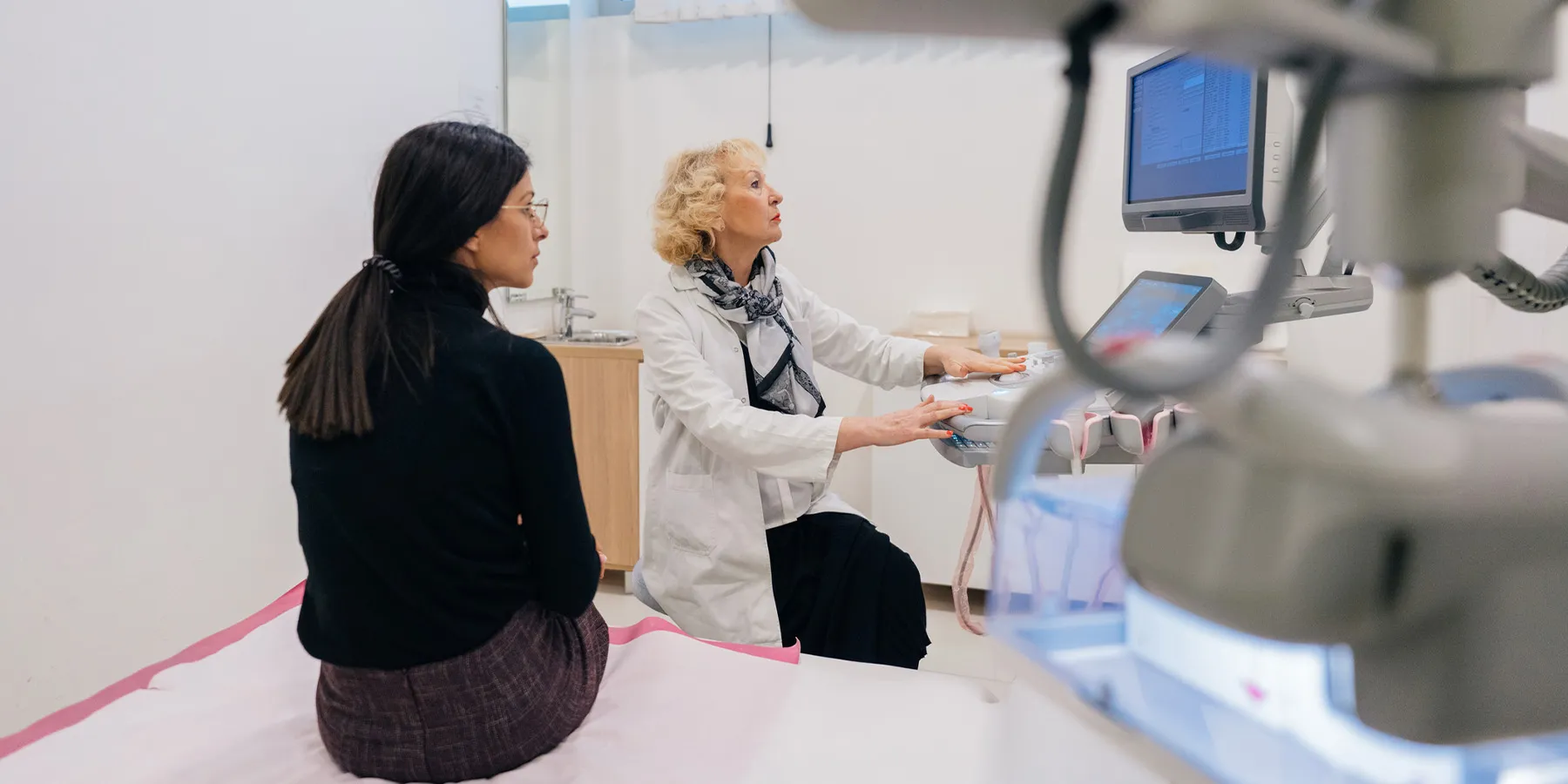 Photo of a woman wearing all black clothes sitting in an exam room with a female doctor wearing a white lab coat.