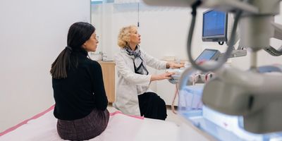 Photo of a woman wearing all black clothes sitting in an exam room with a female doctor wearing a white lab coat.