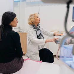 Photo of a woman wearing all black clothes sitting in an exam room with a female doctor wearing a white lab coat.