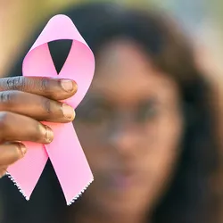 Photo of a Black woman in the background holding up a pink ribbon to represent breast cancer awareness.