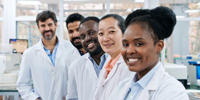 Photo of a diverse group of five laboratorians wearing white lab coats in a clinical laboratory and smiling at the camera.
