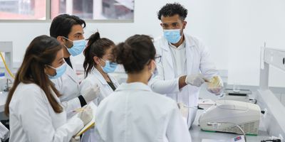 Photo of a group of clinical laboratory professionals wearing white lab coats and blue gloves in the lab and taking notes while learning about clinical lab compliance.