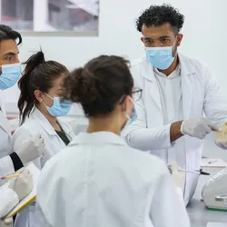 Photo of a group of clinical laboratory professionals wearing white lab coats and blue gloves in the lab and taking notes while learning about clinical lab compliance.