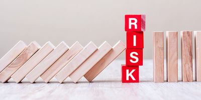 Photo of red wooden blocks stacked on a table with the word “RISK” spelled on the front of them as falling blocks threaten to topple the red blocks.