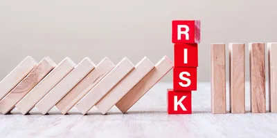 Photo of red wooden blocks stacked on a table with the word “RISK” spelled on the front of them as falling blocks threaten to topple the red blocks.