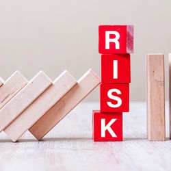 Photo of red wooden blocks stacked on a table with the word “RISK” spelled on the front of them as falling blocks threaten to topple the red blocks.