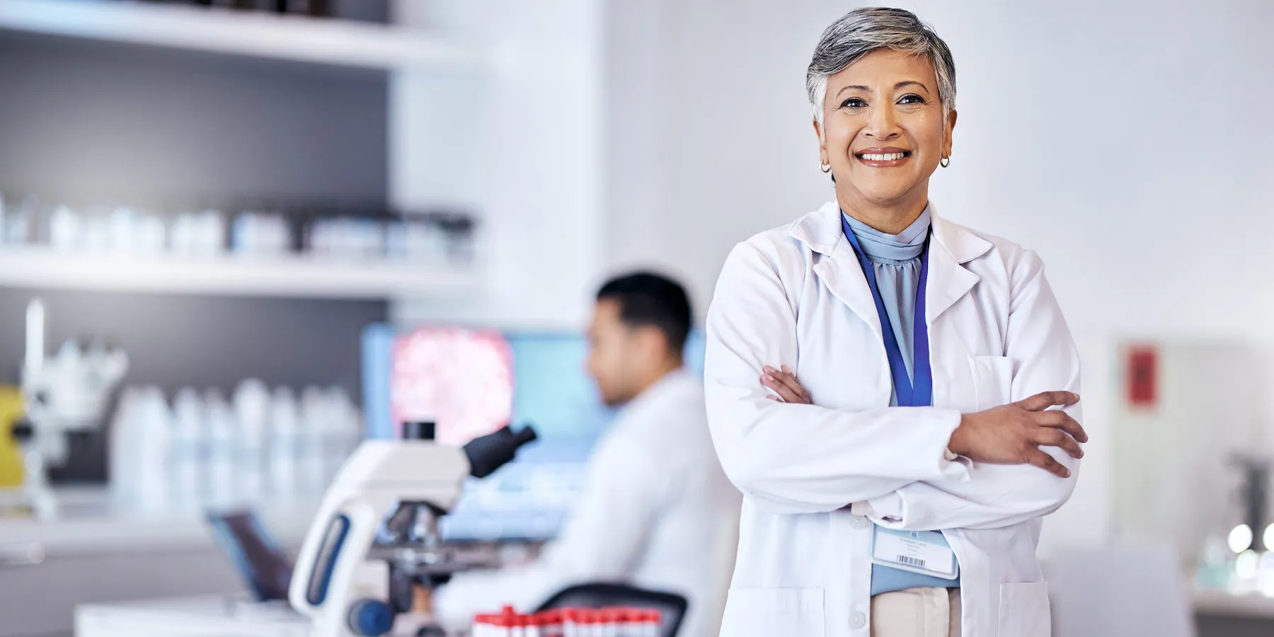  Photo of a clinical laboratory scientist wearing a white lab coat standing with their arms crossed inside of a medical laboratory where testing is conducted.
