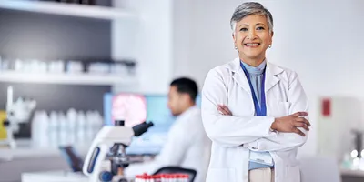 Photo of a clinical laboratory scientist wearing a white lab coat standing with their arms crossed inside of a medical laboratory where testing is conducted.