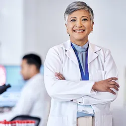 Photo of a clinical laboratory scientist wearing a white lab coat standing with their arms crossed inside of a medical laboratory where testing is conducted.