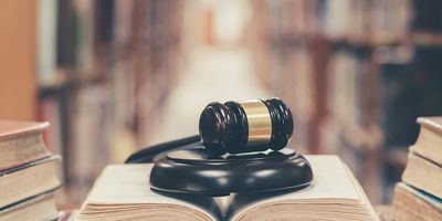 Photo of a book open on a library table with a gavel on top of it with book stacks in the background.