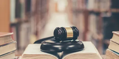Photo of a book open on a library table with a gavel on top of it with book stacks in the background.