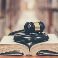 Photo of a book open on a library table with a gavel on top of it with book stacks in the background.