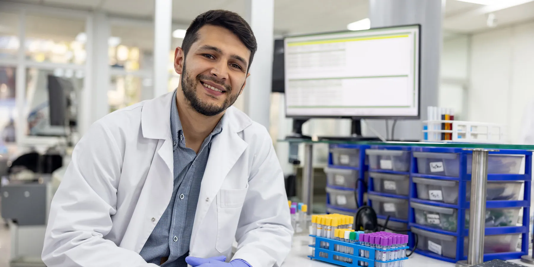 Photo of a male clinical laboratory professional wearing a white lab coat and smiling while sitting in front of a lab bench with a computer that has a laboratory information management system or LIMS.