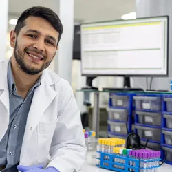 Photo of a male clinical laboratory professional wearing a white lab coat and smiling while sitting in front of a lab bench with a computer that has a laboratory information management system or LIMS.
