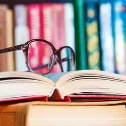 Photo of a pair of black eye glasses on top of an open book inside of a library.