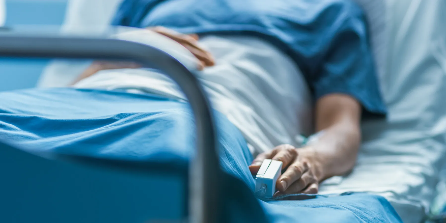 Photo of a clinical trial participant lying in a hospital bed covered with a sheet and wearing an oximeter on their index finger.